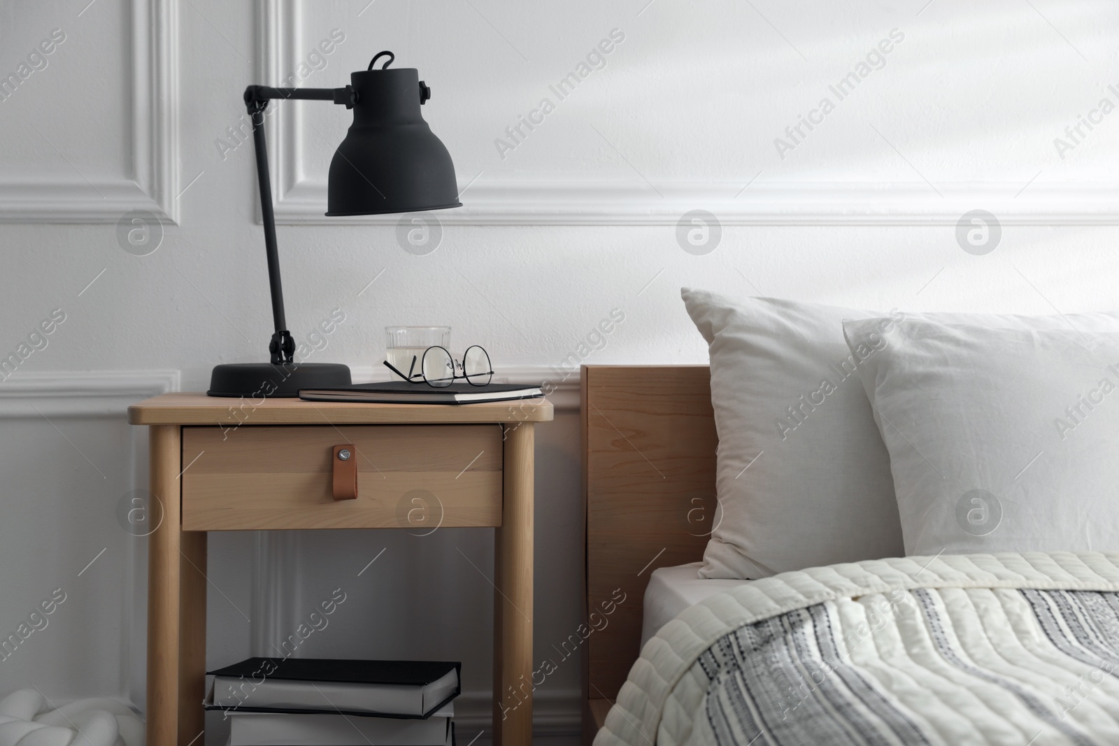 Photo of Bedside table with lamp, notebook, glasses, books and glass of water near bed indoors