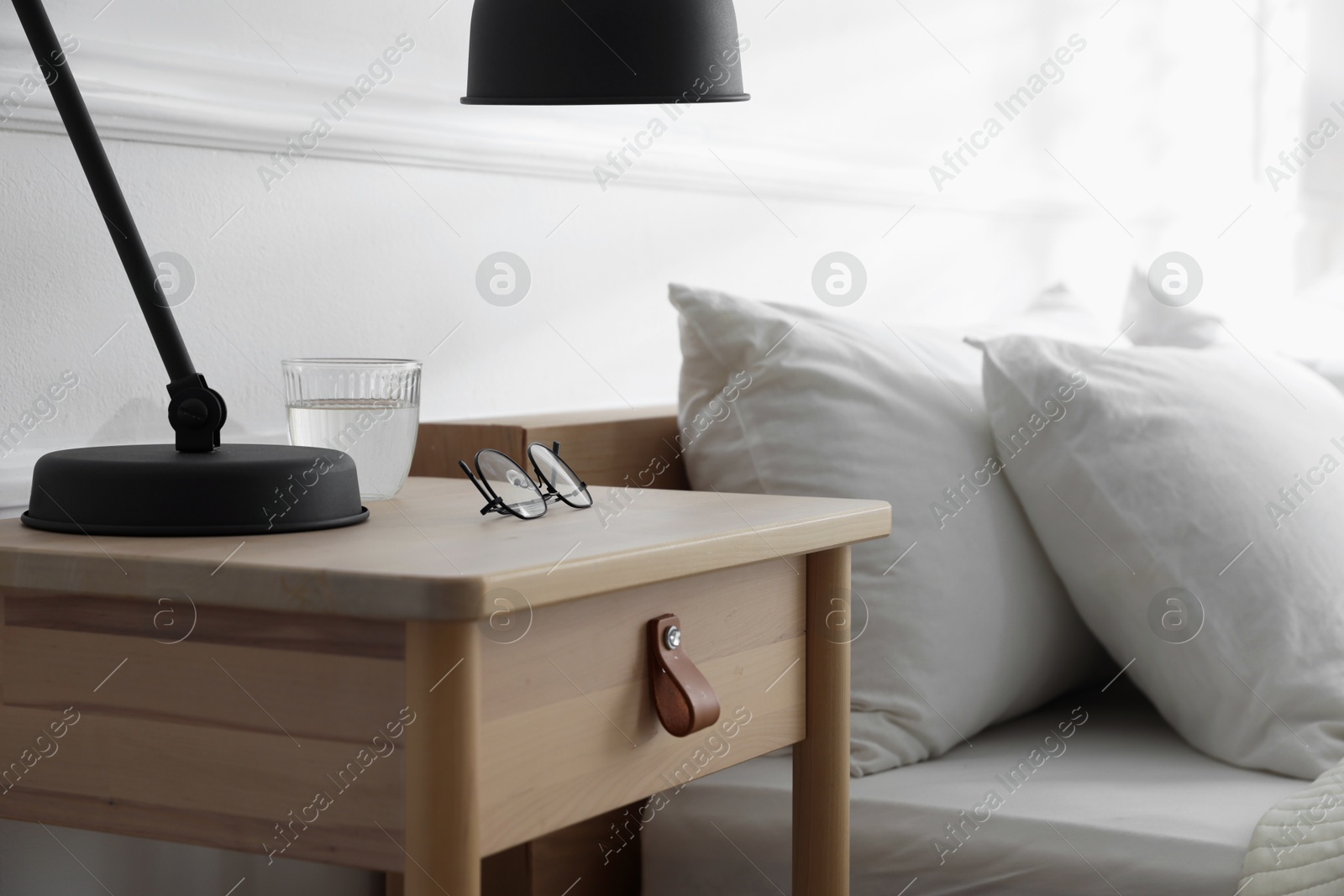 Photo of Bedside table with lamp, notebook, glasses and glass of water near bed indoors, closeup