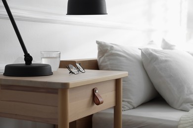 Photo of Bedside table with lamp, notebook, glasses and glass of water near bed indoors, closeup