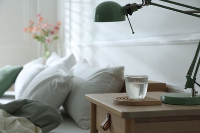 Photo of Bedside table with lamp and glass of water near bed indoors, closeup