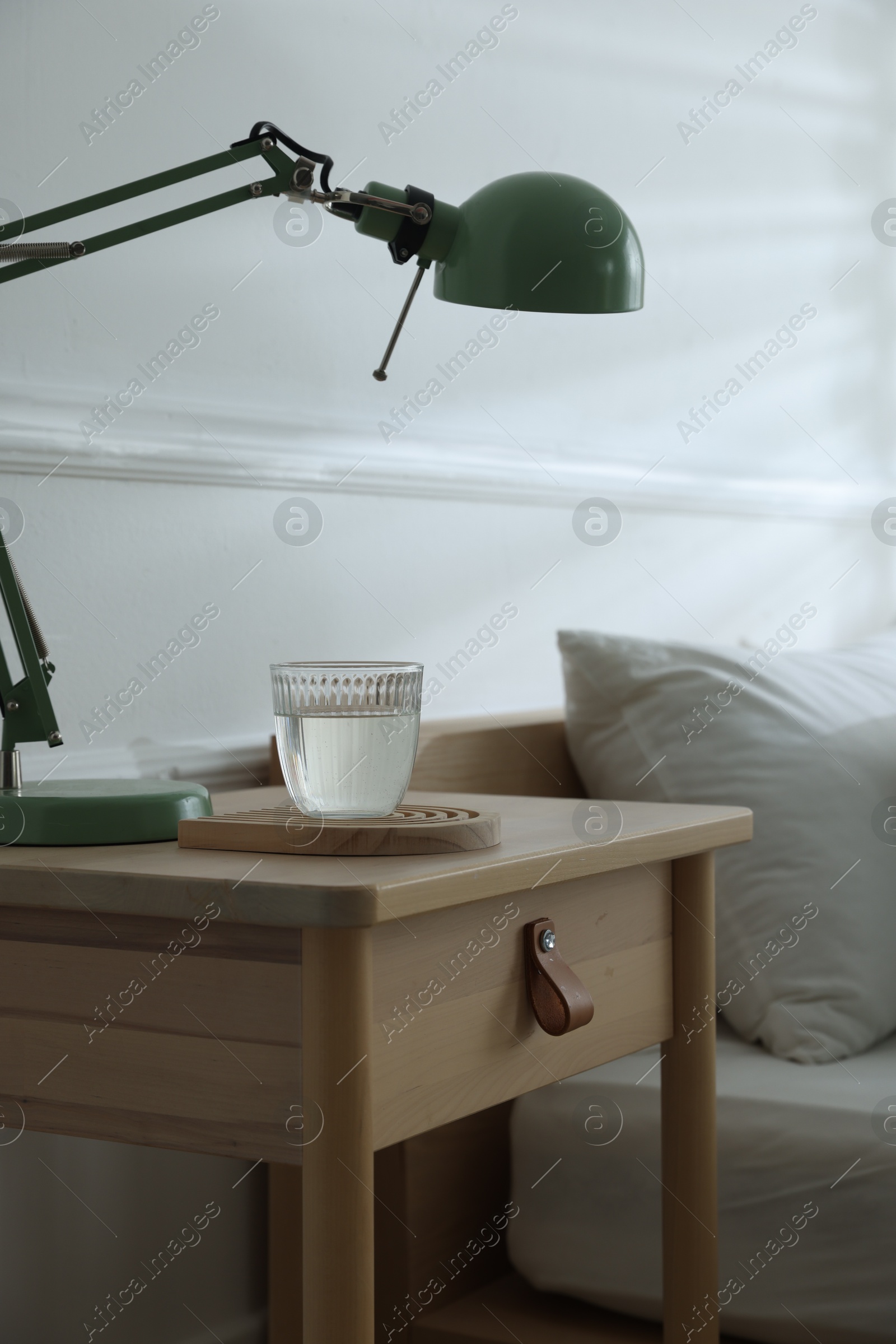 Photo of Bedside table with lamp and glass of water near bed indoors, closeup