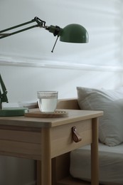 Photo of Bedside table with lamp and glass of water near bed indoors, closeup