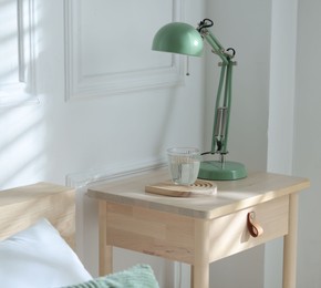 Photo of Bedside table with lamp and glass of water near bed indoors