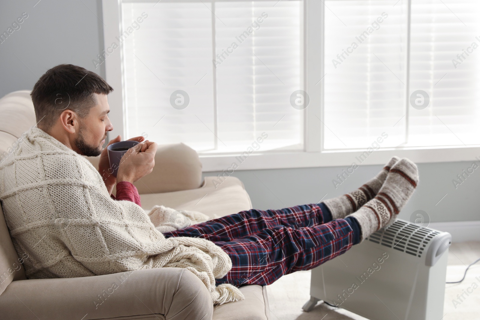 Photo of Man with cup of hot drink warming feet on electric heater at home. Space for text