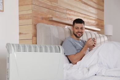 Photo of Man with cup of hot drink in bed near electric heater at home