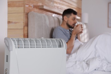 Photo of Man with cup of hot drink in bed at home, focus on electric heater