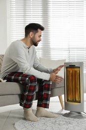 Photo of Young man warming hands near electric heater at home