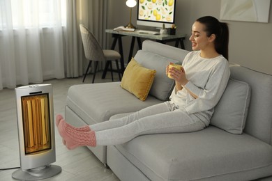 Photo of Young woman with cup of drink sitting on sofa near electric heater at home