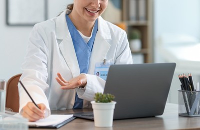 Photo of Telemedicine. Doctor making notes while having video call with patient via laptop indoors, closeup