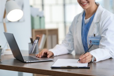 Photo of Telemedicine. Doctor making notes while having video call with patient via laptop indoors, closeup