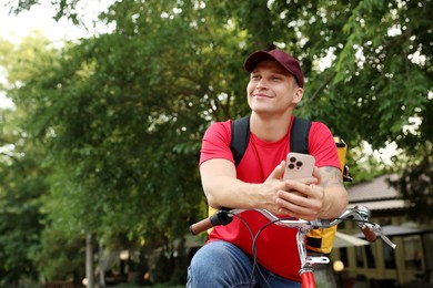 Photo of Courier with thermo bag and smartphone on bicycle outdoors, space for text. Food delivery service