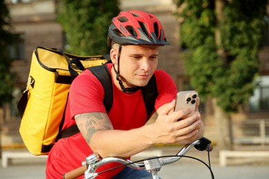 Photo of Courier with thermo bag and smartphone on bicycle outdoors. Food delivery service
