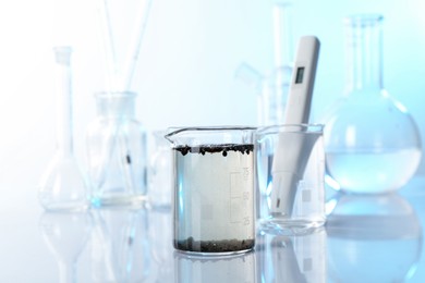 Photo of Glass beaker with dirty water, other glassware and digital pH meter on mirror surface