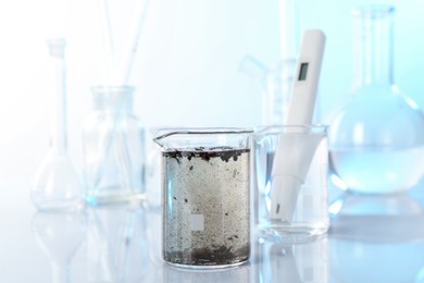 Photo of Glass beaker with dirty water, other glassware and digital pH meter on mirror surface, closeup