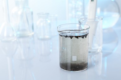 Photo of Glass beaker with dirty water, other glassware and digital pH meter on mirror surface, closeup. Space for text