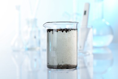 Photo of Glass beaker with dirty water, other glassware and digital pH meter on mirror surface, closeup