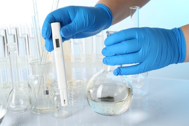 Photo of Examination of water quality. Scientist with flask of sample and digital pH meter at table against light background, closeup