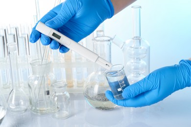 Photo of Examination of water quality. Scientist with flask of sample and digital pH meter at table against light background, closeup