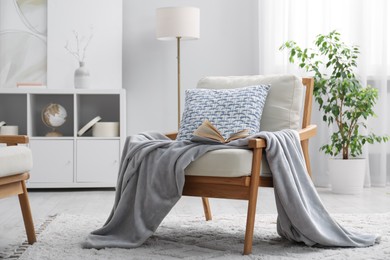 Photo of Comfortable armchair with book, pillow and blanket in living room. Interior design