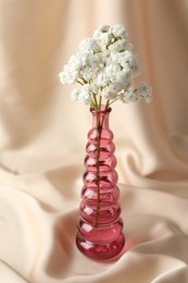 Photo of Beautiful gypsophila flowers in glass vase on beige cloth, closeup