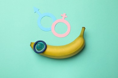 Photo of Sex education. Banana with condom, paper female and male gender symbols on turquoise background, flat lay