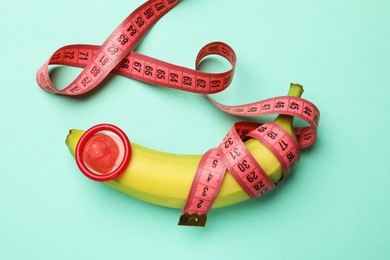 Photo of Sex education. Banana with condom and measuring tape on turquoise background, top view