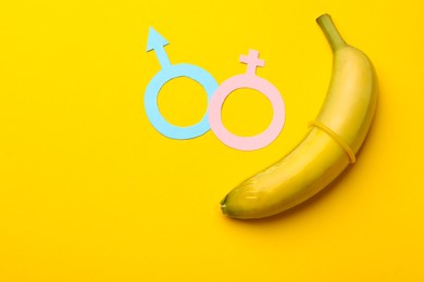 Photo of Sex education. Banana with condom, paper female and male gender symbols on yellow background, flat lay