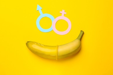 Photo of Sex education. Banana with condom, paper female and male gender symbols on yellow background, flat lay