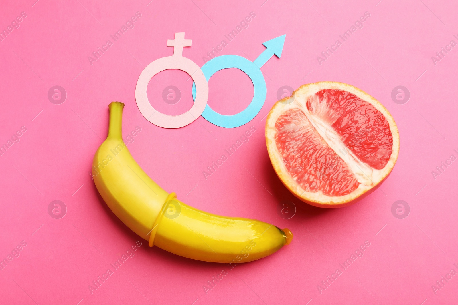 Sex education. Banana with condom, grapefruit, paper female and male gender symbols on pink background, flat lay Photo of Sex education. Banana with condom, grapefruit, paper female and male gender symbols on pink background, flat lay