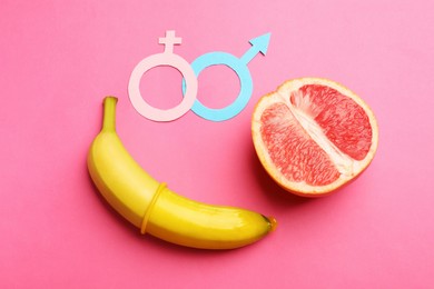 Photo of Sex education. Banana with condom, grapefruit, paper female and male gender symbols on pink background, flat lay