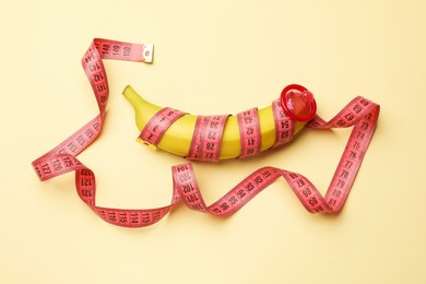 Photo of Sex education. Banana with condom and measuring tape on beige background, top view