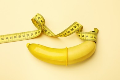 Photo of Sex education. Banana with condom and measuring tape on beige background, top view