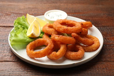 Photo of Fried squid rings, lemon, lettuce and sauce on wooden table, closeup