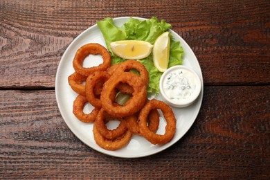 Photo of Fried squid rings, lemon, lettuce and sauce on wooden table, top view