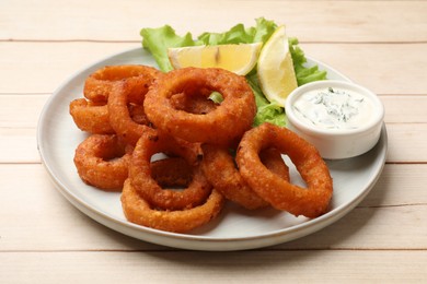 Photo of Fried squid rings, lemon, lettuce and sauce on light wooden table, closeup