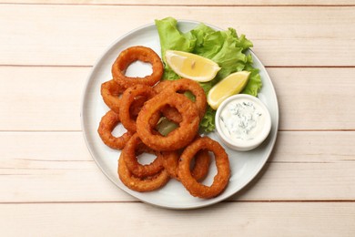 Photo of Fried squid rings, lemon, lettuce and sauce on light wooden table, top view