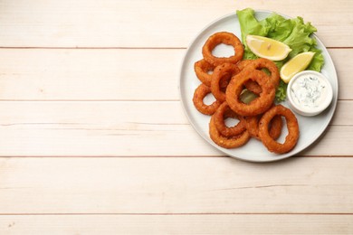 Photo of Fried squid rings, lemon, lettuce and sauce on light wooden table, top view. Space for text