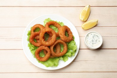 Photo of Fried squid rings, lemon, lettuce and sauce on light wooden table, flat lay