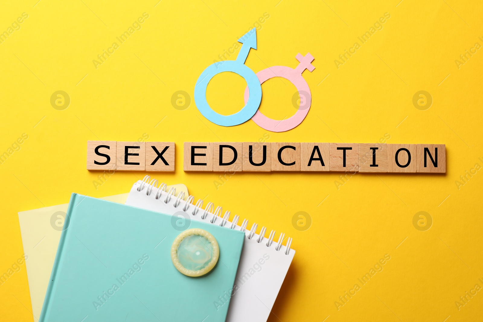 Photo of Words Sex Education made of wooden squares, gender symbols, condom and notebooks on yellow background, flat lay