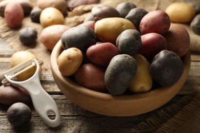 Photo of Different types of potatoes in bowl and peeler on wooden table, closeup