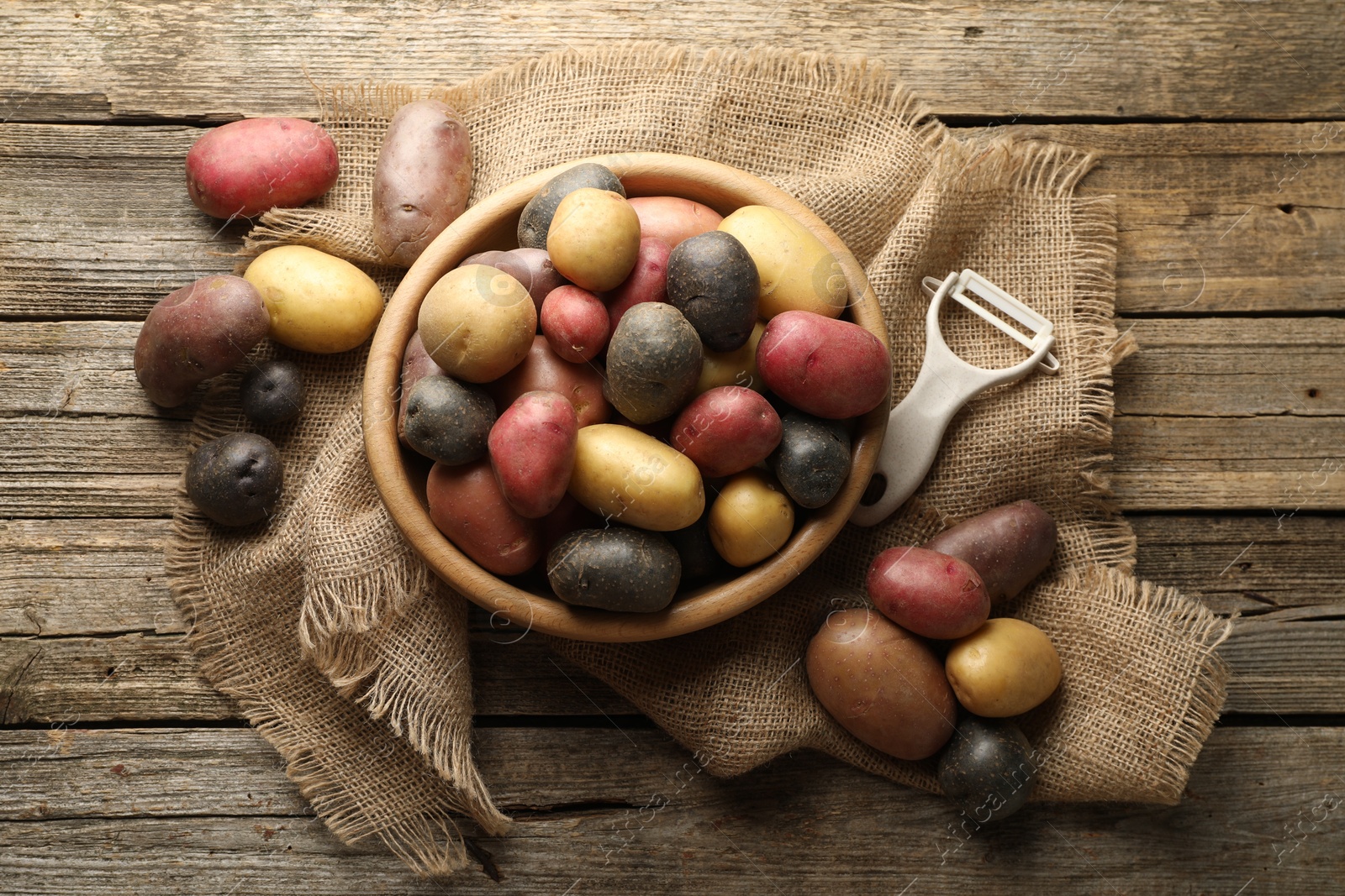 Photo of Different types of potatoes in bowl and peeler on wooden table, flat lay