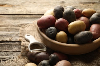 Photo of Different types of potatoes in bowl and peeler on wooden table, closeup. Space for text