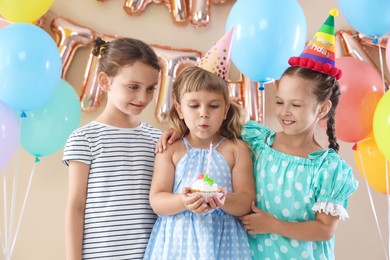 Photo of Birthday celebration. Cute little girl blowing out candle on cupcake, her friends and festive decor against beige background