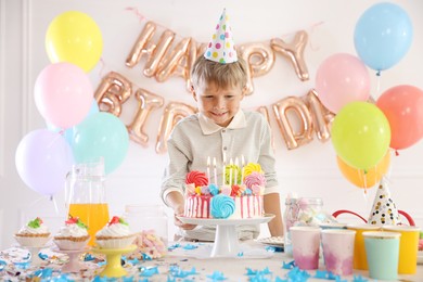 Photo of Birthday celebration. Cute little boy at table with tasty cake and different treats in festive decorated room