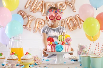 Photo of Birthday celebration. Cute little girl at table with tasty cake and different treats in festive decorated room