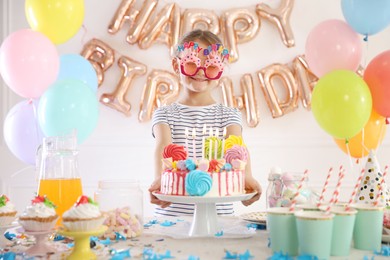 Photo of Birthday celebration. Cute little girl at table with tasty cake and different treats in festive decorated room