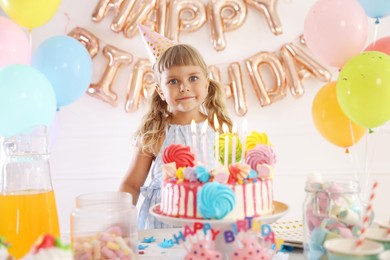 Photo of Birthday celebration. Cute little girl at table with tasty cake and different treats in festive decorated room