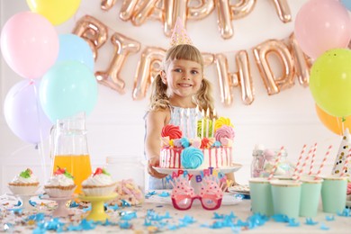 Photo of Birthday celebration. Cute little girl at table with tasty cake and different treats in festive decorated room
