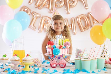 Photo of Birthday celebration. Cute little girl at table with tasty cake and different treats in festive decorated room