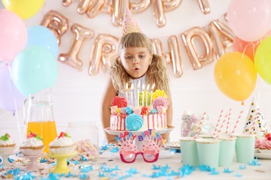 Photo of Birthday celebration. Cute little girl blowing out candles on cake at table in festive decorated room